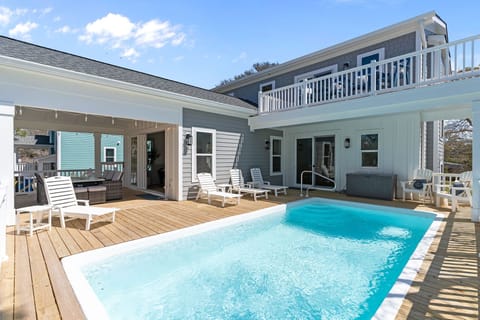Bright private pool with clear blue water on a wood deck. White lounge chairs surround the pool. Covered patio with outdoor seating, sliding glass doors, gray coastal siding, balcony above, and sunny blue sky at this Oak Island beach rental.
