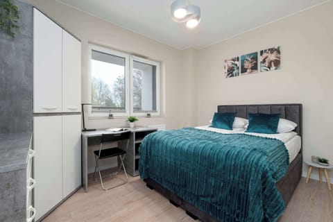 A brightly lit bedroom features a double bed with a teal throw and matching pillows. A small workstation with a desk and chair is tucked neatly into the corner by the window.