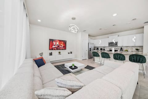 Step into a bright living area with a spacious light-colored sectional sofa adorned with vibrant pillows. The sleek kitchen, equipped with stylish bar stools, invites lively gatherings, all enhanced by warm, cozy lighting.