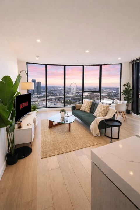 Curved living room with a green sofa, glass coffee table, and Netflix-ready TV, all facing a sweeping sunset view of the Melbourne Star observation wheel and city skyline through floor-to-ceiling windows.