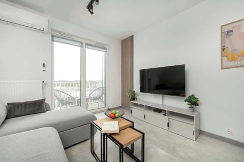 A bright living room features a grey L-shaped sofa and a wall-mounted television. Large glass doors lead out to a private balcony, filling the space with natural light.