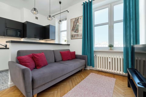 A modern living room features a grey sofa accented with vibrant red pillows. Two large windows with teal curtains allow natural light to fill the space, highlighting the classic herringbone wooden floor.