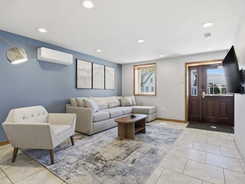 The living room features a light-gray chaise sectional, upholstered armchair, oval wood coffee table, patterned rug, wall-mounted TV, mini-split, windows, recessed lighting, and double entry doors.