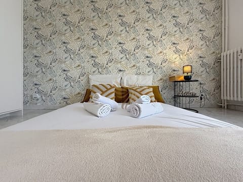 Réveillez-vous dans ce cocon végétal : lit bas habillé de coussins moutarde, papier peint feuillagé apaisant et lampe tamisée sur la table `dream`, une chambre cosy pour une nuit reposante.