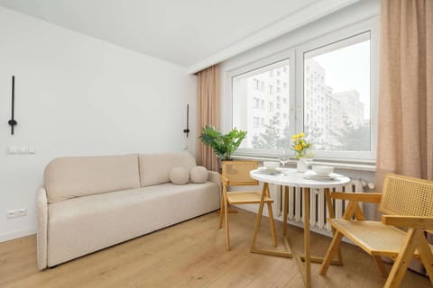 A compact living area with a sofa positioned next to a small dining setup. A TV unit stands opposite the seating area, making it ideal for relaxation. Neutral tones and simple furnishings create a calm, modern atmosphere.