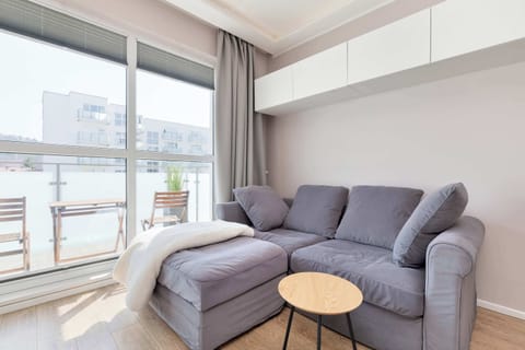 A light-filled living area features a grey corner sofa adorned with white blankets and a small wooden side table.