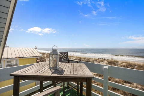 Wide ocean view across sandy dunes and sea oats to the shoreline, with beachgoers, rolling waves, and a bright blue sky stretching over the Atlantic.