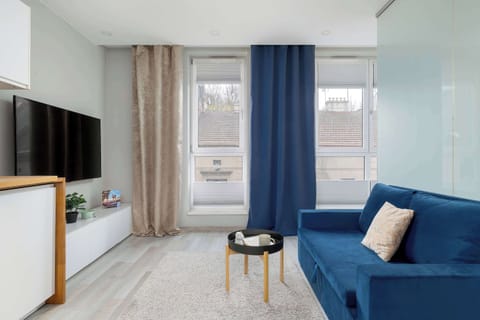 A bright living area features a vibrant navy blue sofa positioned across from a minimalist white media console and television. Natural light streams in from large windows dressed with navy blue curtains.
