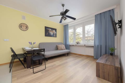 A spacious living area contains a comfortable-looking grey sofa and a flat-screen television. Two large windows allow plenty of natural light into the room, which also features wooden flooring.