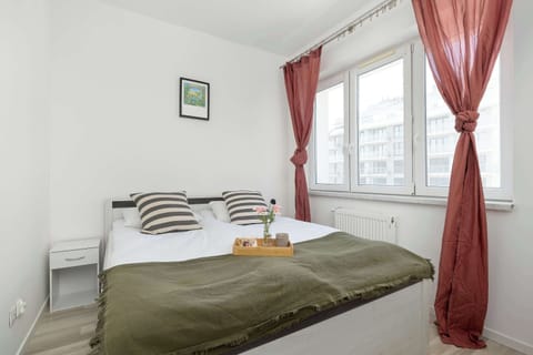 A bright bedroom features a large double bed with a green throw and decorative pillows. A small white nightstand sits next to the bed under a window with reddish curtains.
