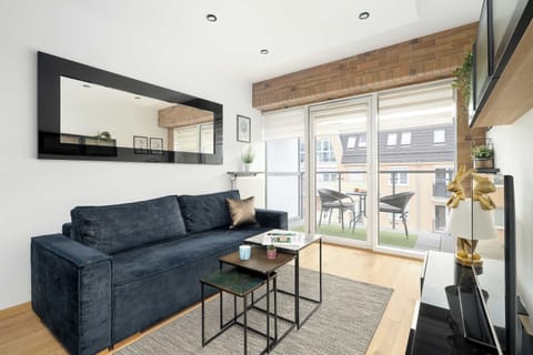 A modern living room features a dark blue sofa and a minimalist coffee table, brightened by large floor-to-ceiling windows leading to a balcony. The space is accented by a decorative brick wall and light wooden flooring.