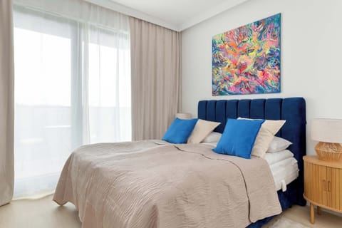 A bright bedroom features a navy blue upholstered headboard and a colorful abstract painting on the wall. The bed is neatly made with a beige quilted spread and accent pillows in deep blue and tan.