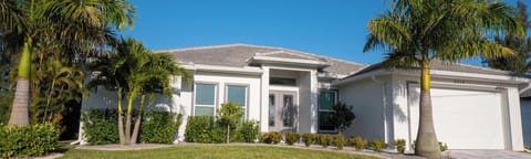 Single-story white villa with gray tile roof, flanked by tall palm trees and neatly trimmed green shrubs.