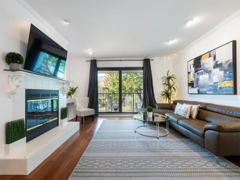 Living room features a dark leather sectional, glass coffee table, and a wall-mounted TV above a white tiled fireplace.