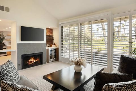 Living room with tile-surround fireplace, wall-mounted TV, wine fridge bar nook, and plantation shutters framing the golf course