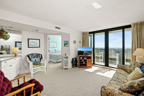 Relax in your spacious living room with comfortable seating, entertainment center, and stunning balcony views through floor-to-ceiling windows.