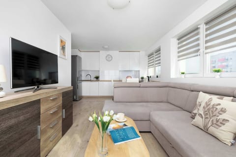 A bright living room features a large grey corner sofa and a wooden media console under a wall-mounted TV.