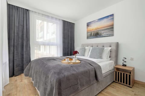 A bright bedroom features a plush grey tufted bed with a matching quilted throw and a sunset-themed landscape painting on the wall. A simple wooden bedside table adds a natural touch to the clean, white interior.