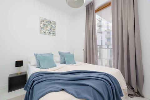 A bright bedroom featuring a double bed with blue bedding, soft curtains, and natural light coming through a tall window.