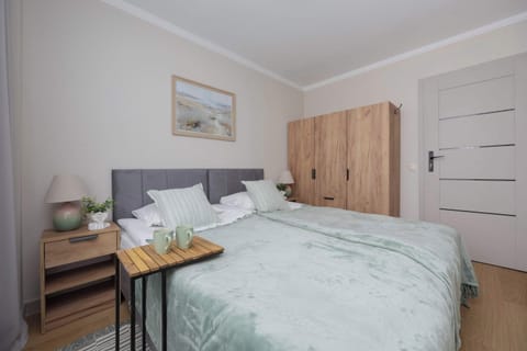 A cozy bedroom features a bed with light green bedding and a wooden wardrobe. A small wooden side table sits next to the bed under a minimalist framed picture on the wall.