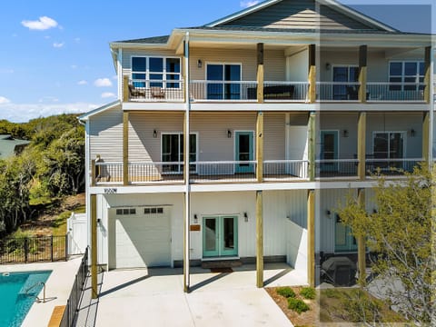 Modern three-story coastal home with multiple balconies and pool, surrounded by lush natural landscape under blue skies.