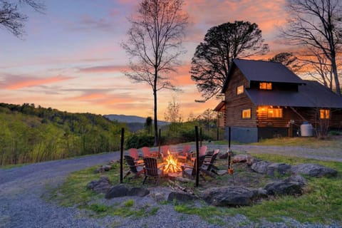 Hillside cabin with a glowing fire pit and Adirondack chairs, set against a colorful mountain sunset.