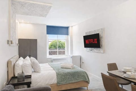 Bright and stylish bedroom featuring a plush double bed with luxury linens, feature wallpaper and soft lighting. Includes a cosy sofa, Smart TV, wardrobe and dining space, with large windows bringing in natural light. Perfect for relaxing stays.