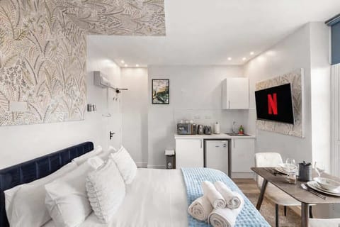 right and stylish bedroom featuring a plush double bed with luxury linens, feature wallpaper and soft lighting. Includes a  Smart TV, wardrobe and dining space, with large windows bringing in natural light. Perfect for relaxing stays.