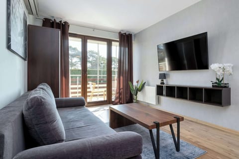A cozy lounge area featuring a gray couch facing a wall-mounted TV. Large balcony doors bring in daylight and open up the space.
