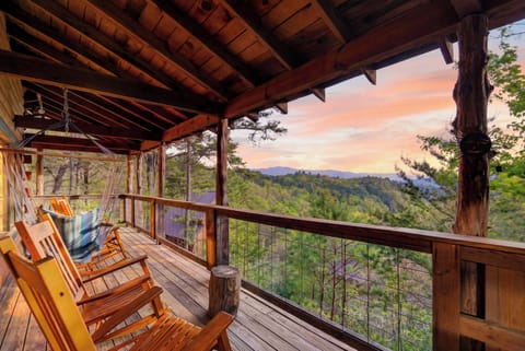 Sunset‑lit cabin deck with rocking chairs and a hammock swing overlooking peaceful forested mountains.