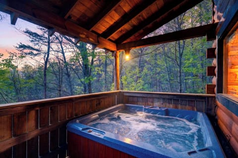 Covered cabin deck with a bubbling hot tub, warm evening light, and peaceful forest views at dusk.