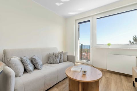 A light-filled living area has a pale-grey fabric sofa adorned with textured patterned and beige pillows. A low, round wooden coffee table stands on the light wood floor, and large windows show a view of trees and buildings.