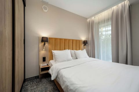 A modern hotel bedroom with a large bed and soft lighting. The space feels warm and inviting with neutral tones and curtains by the window.