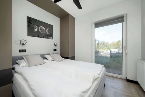A bright bedroom features a large double bed with white linens and a minimalist wall decoration above the headboard. A large glass door leads directly to the garden, filling the room with natural light.