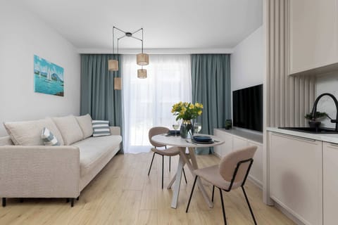 A bright studio apartment with a small dining table, sofa, and kitchenette arranged in a clean, modern layout. Light wood flooring and soft colors create an airy atmosphere.