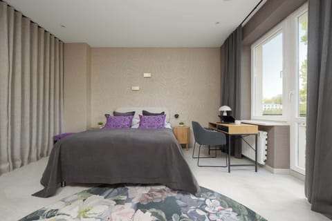 A cozy sleeping area featuring a neatly made bed with soft gray linens and purple cushions. A small desk stands by the window, creating a functional corner for work or getting ready.