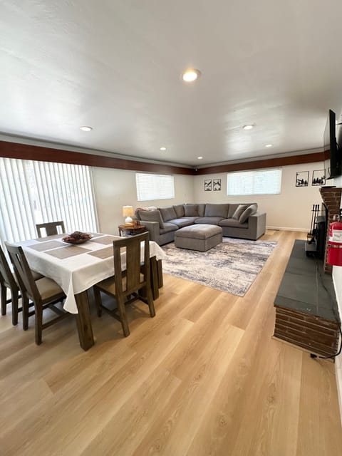 Living room with sectional sofa, ottoman, fireplace, flat-screen TV, dining table seating, sliding door, windows, and table lamp.