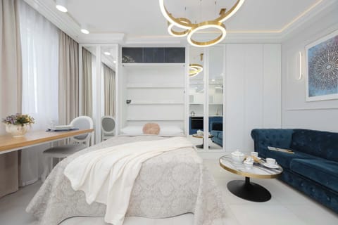 A bright bedroom features a plush bed with a cream-colored throw and a deep blue velvet sofa positioned along the side wall. Large floor-to-ceiling mirrors reflect the light, enhancing the spacious feel of the modern interior.