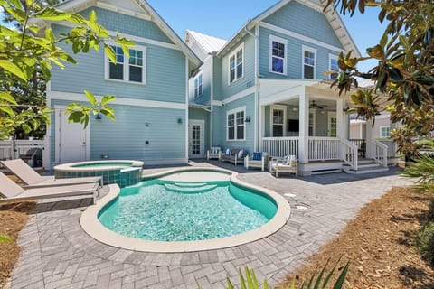 This coastal-style home features a sparkling pool, hot tub, and covered porch with ample seating—ideal for relaxing under sunny skies surrounded by lush tropical greenery and beachside charm.