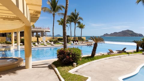 Curved yellow columns frame the blue pool beside palm trees and green shrubbery with ocean and island views.