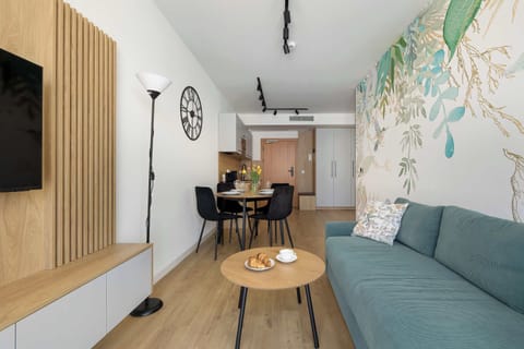 Living and dining space, combining the green sofa, round table, and modern chairs. The floral wall design and light streaming through the windows give the room a fresh and welcoming character.