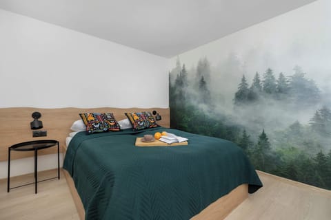 A cozy bedroom features a dark green duvet on a double bed, accented by a large forest-mist mural on the headboard wall.
