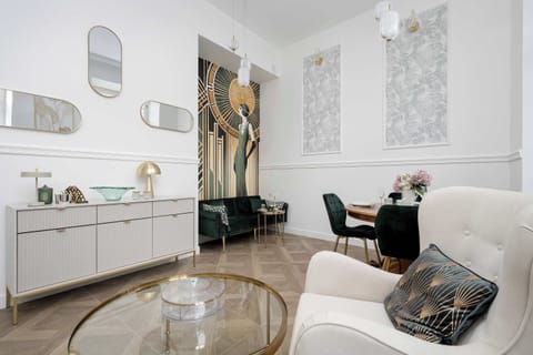 An elegant living area is decorated in an Art Deco style, featuring a white sideboard with gold accents and a comfortable cream armchair. The herringbone wood floors and high ceilings create a spacious, luxury feel.