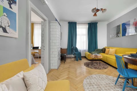 A spacious living room featuring a striking yellow corner sofa set against a light gray wall adorned with modern artwork. In the background, you can see the passageway to the bedroom and a table with blue chairs.