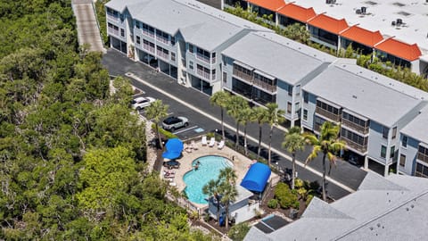 Aerial view of the resort property showing multi-story buildings with balconies, a central swimming pool with blue umbrellas, and lush tropical landscaping.