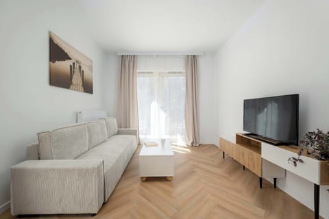 A living room with a comfortable sofa, a TV on a low stand, and a wooden floor arranged in a herringbone pattern. The space feels warm and inviting, ideal for relaxing after a long day.