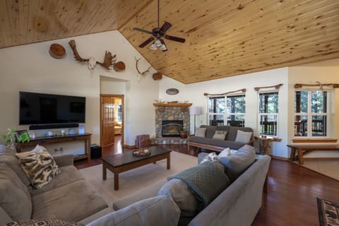Settle into this inviting living space where pine ceilings, a cozy stone fireplace, and rustic charm create your perfect mountain retreat. Large windows frame serene forest views while you relax.