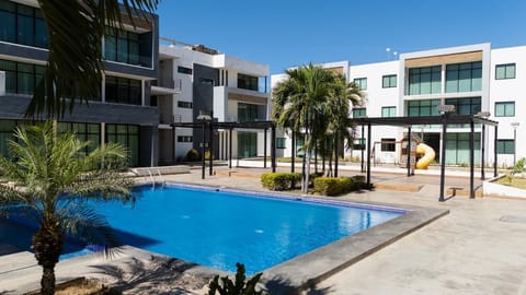 Swimming pool surrounded by concrete deck, palm trees, and black metal pergolas under clear blue sky.