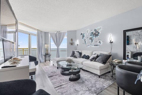 Stylish oceanfront living room on the 18th floor featuring luxurious seating, plush couch, upholstered chairs, Smart TV, indoor electric fireplace, floor-to-ceiling windows, and private balcony access with breathtaking coastal views.