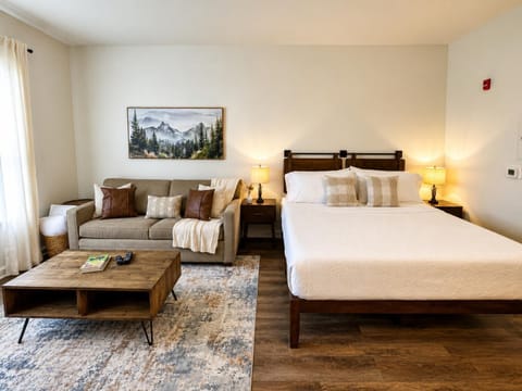 Wood-framed king bed and beige sofa flank a rustic coffee table on a patterned area rug in the studio.